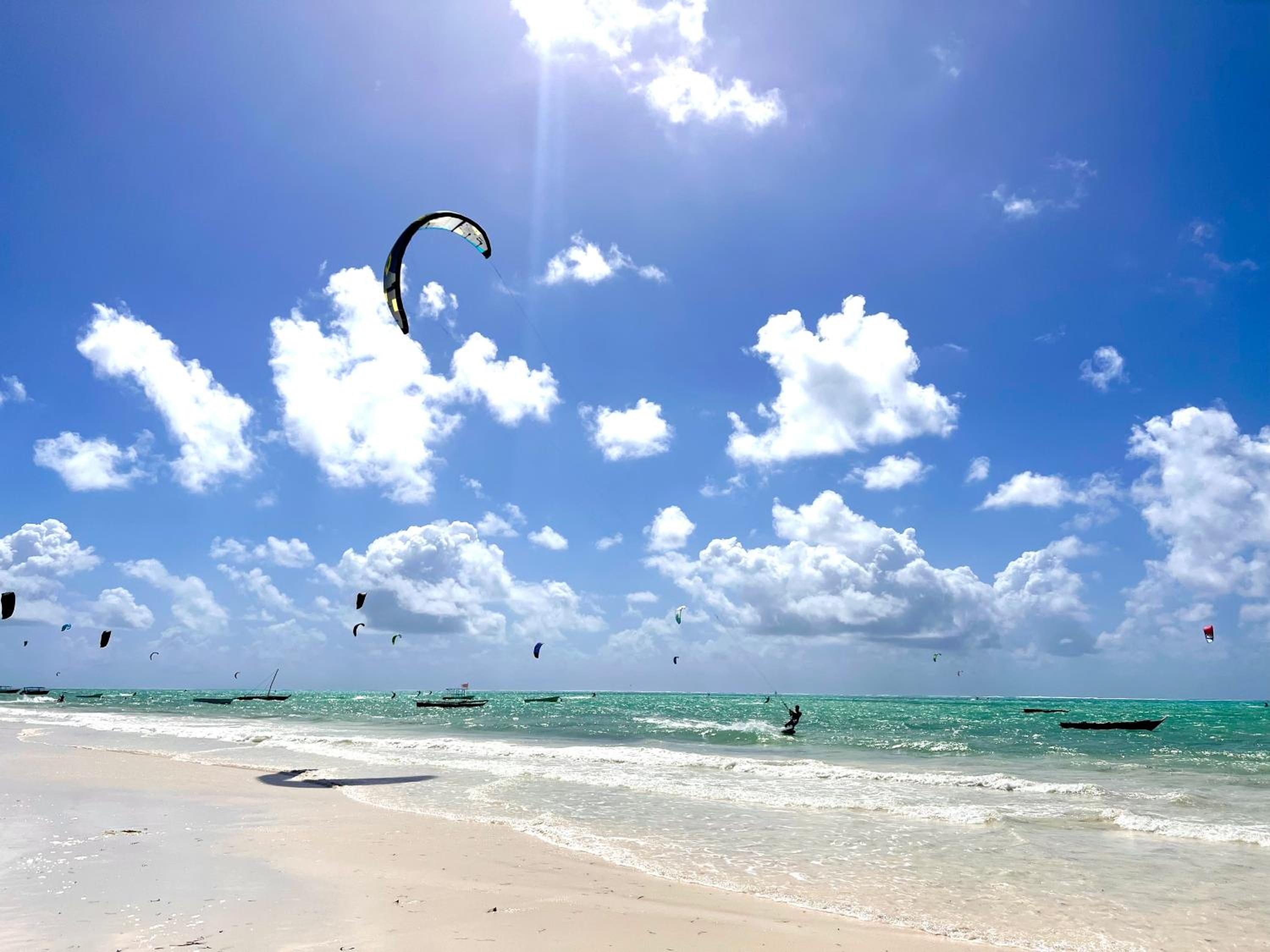 Paje Beach under a blue sky full of clouds: white sand, turquoise water and kites over the lagoon.