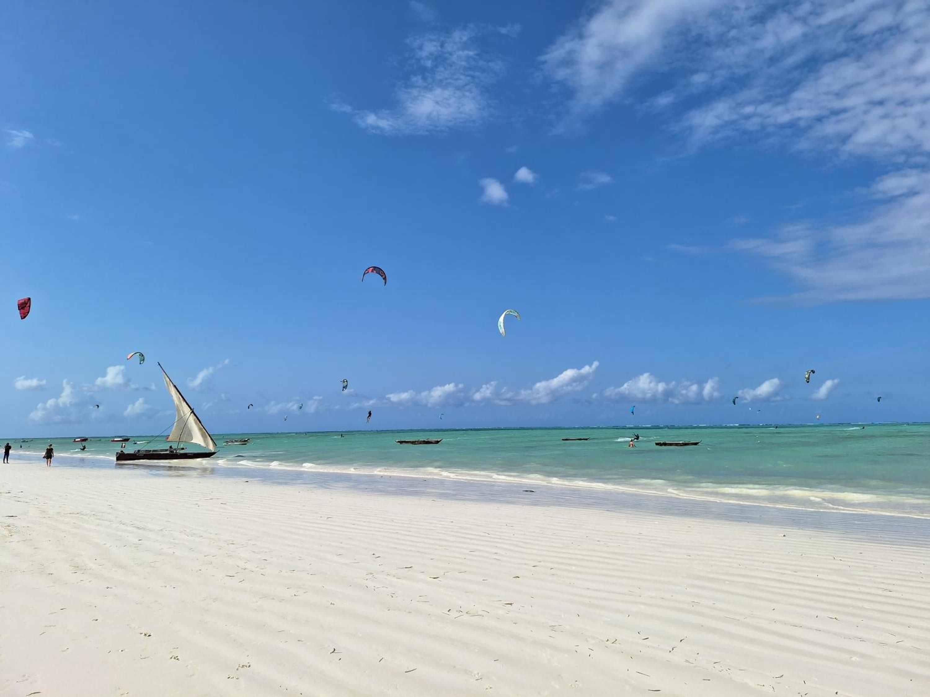 A traditional dhow sailing boat pulled up on the white sand, kites in the sky beyond.
