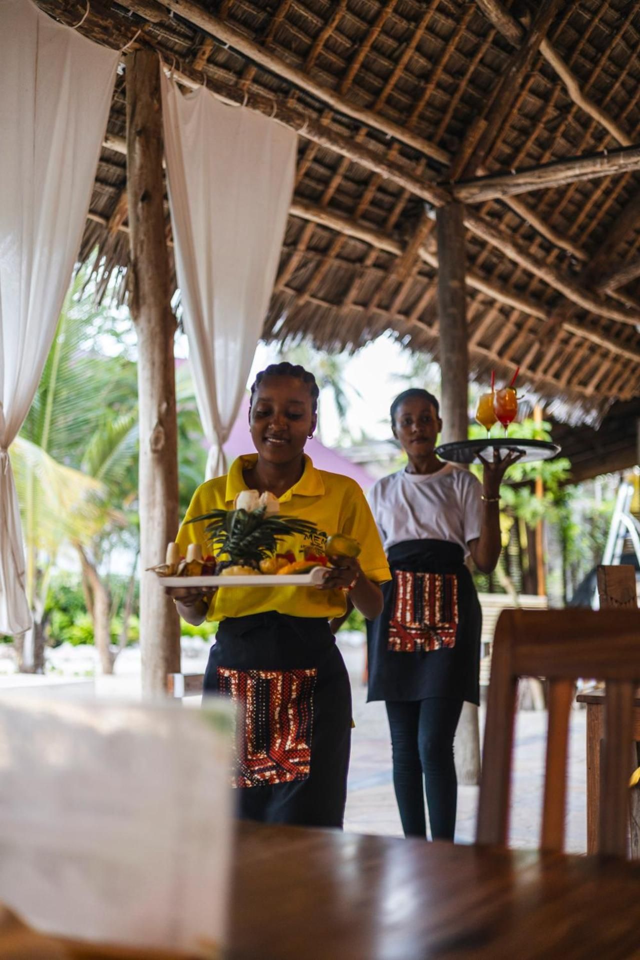 A waitress in a yellow uniform carrying a fruit platter, followed by a colleague with a tray of drinks, under the thatched restaurant roof.