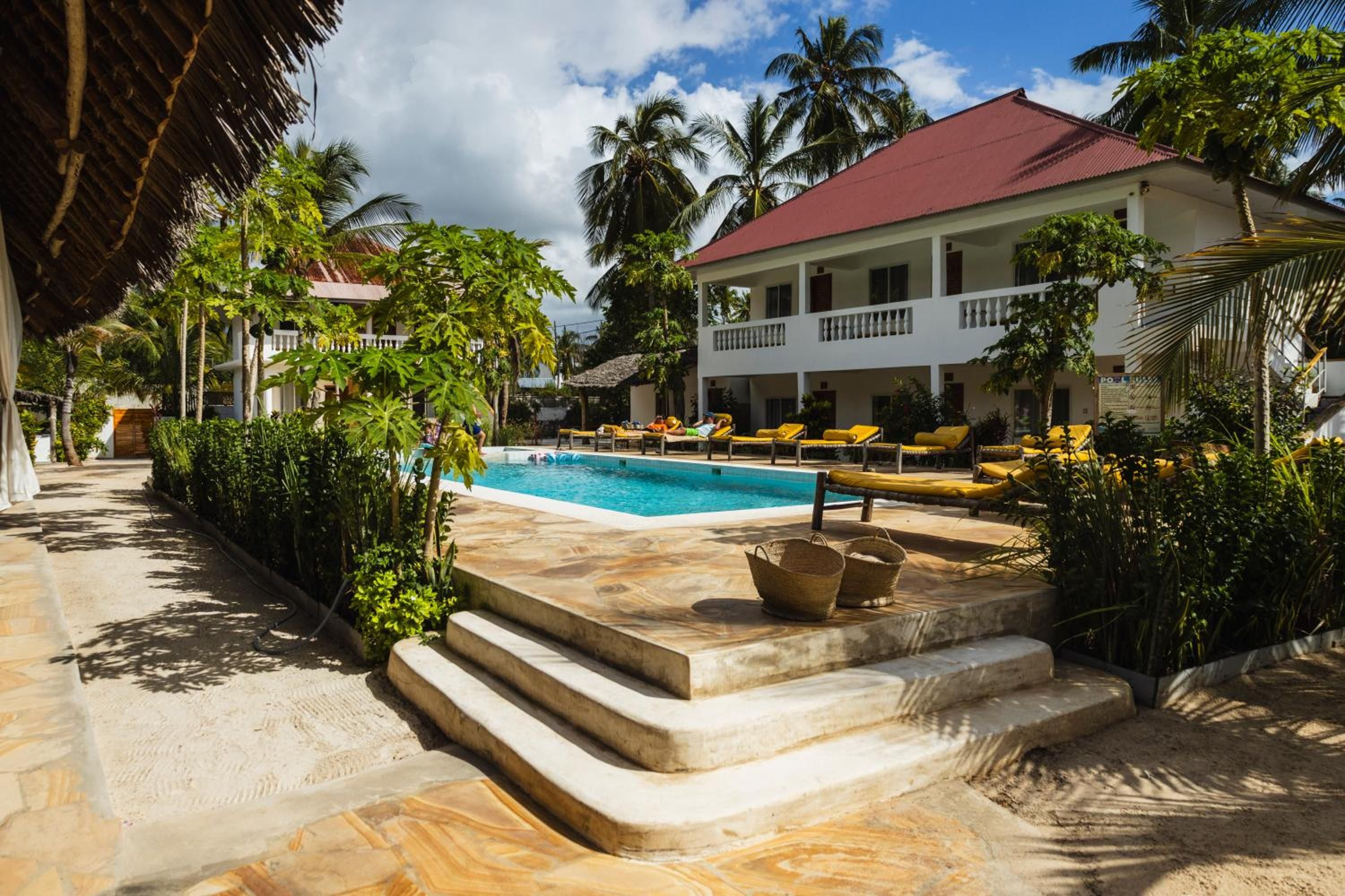 The pool on a sunny day: stone steps lead down to the water, with loungers, palms and the white two-storey building behind.