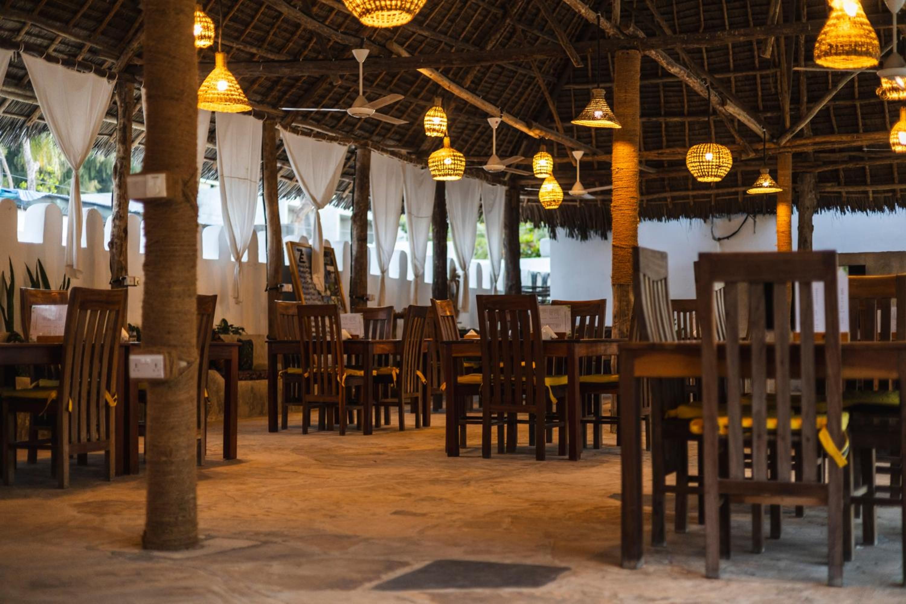 The restaurant in the evening: wooden tables and chairs under a high thatched roof, lit by woven pendant lamps.