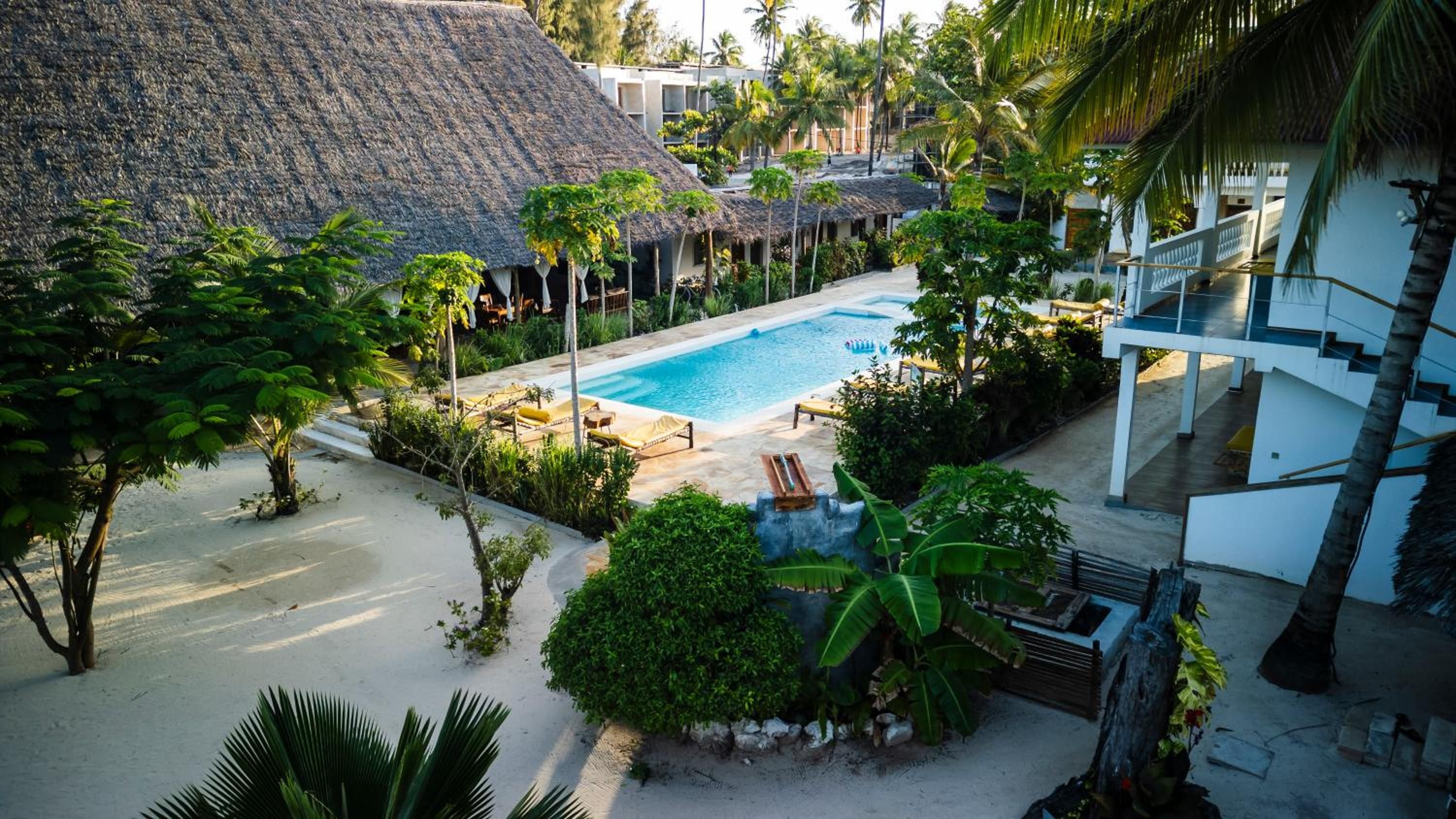 Mem at golden hour: the pool among palms and makuti-thatched pavilions, with the white two-storey rooms on the right.