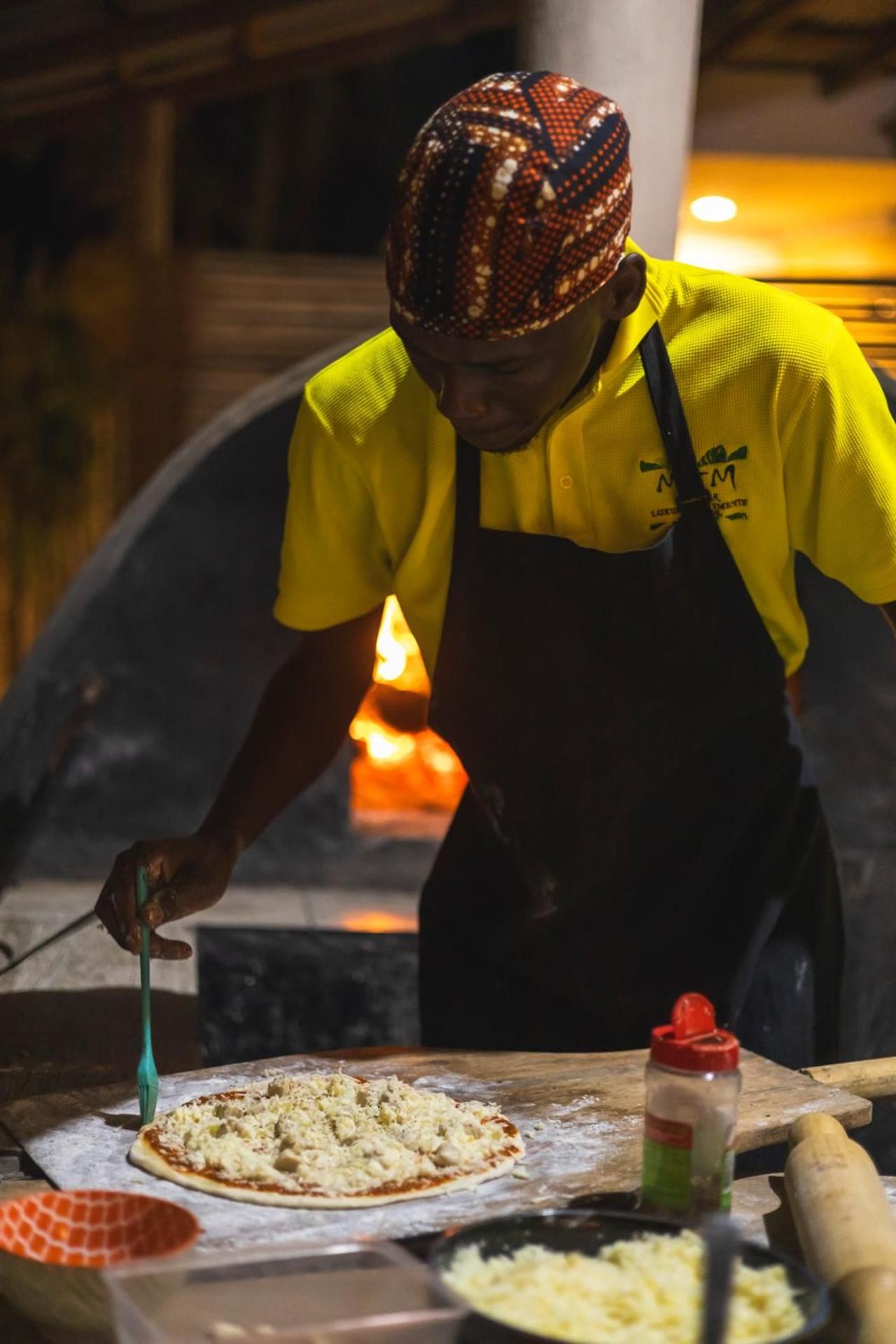 A cook in a yellow shirt and patterned cap topping a pizza by the glowing wood-fired oven.